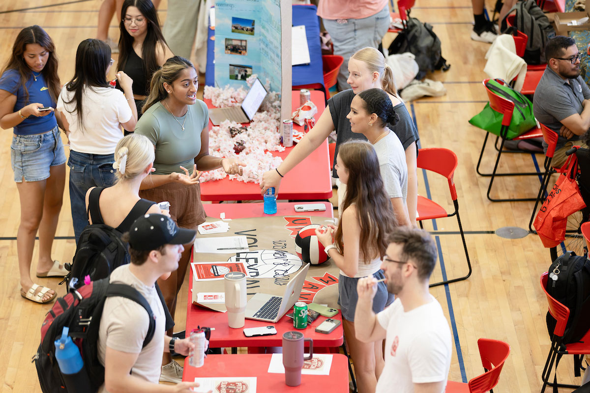 Students gather around a table at a campus involvement fair, talking with representatives and looking at event materials, a laptop, and sign-up sheets.
