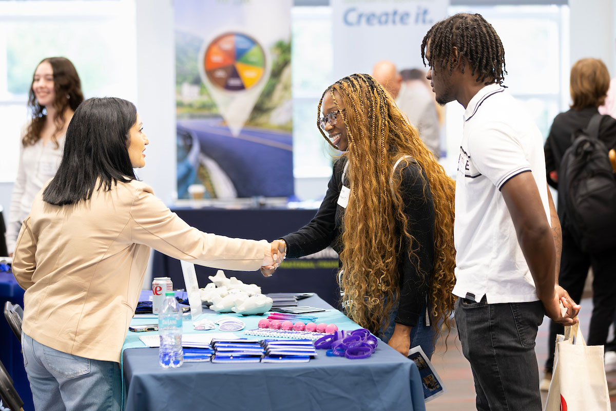Two students shake hand with an employer at a career fair table.