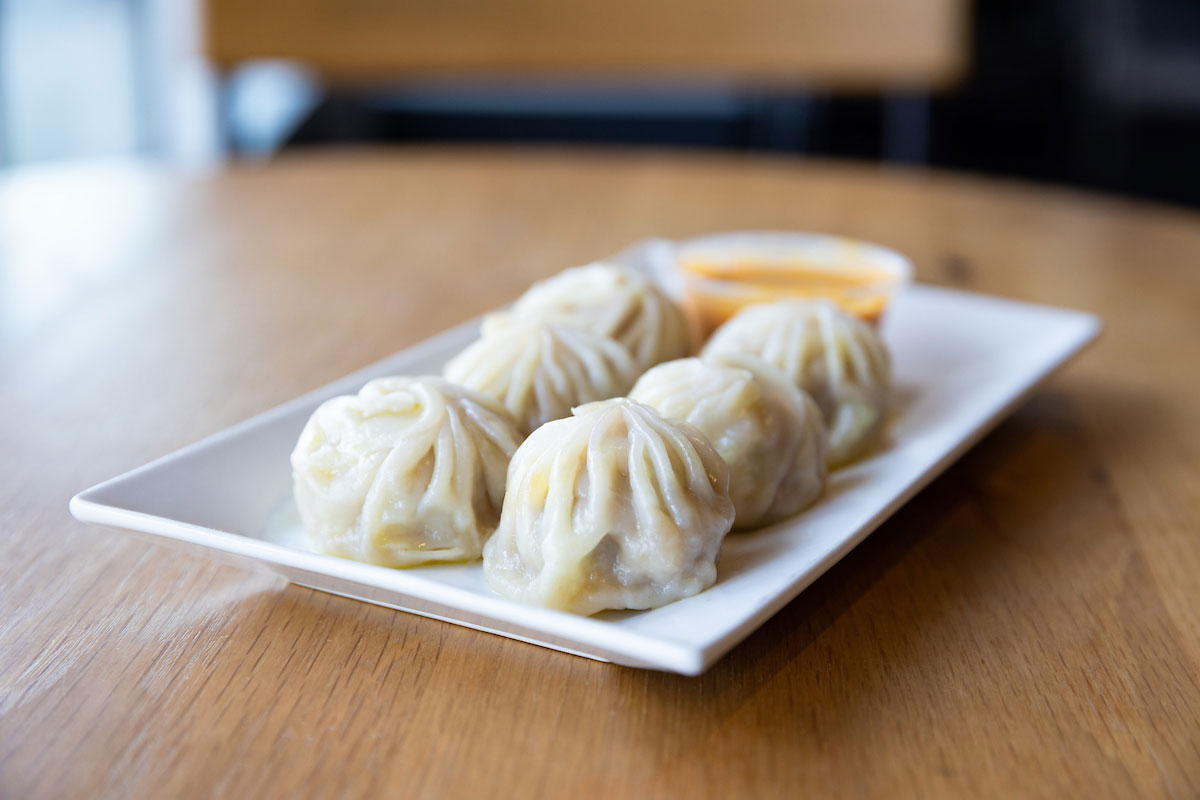 Six dumplings sit on a plate with a small bowl of sauce.