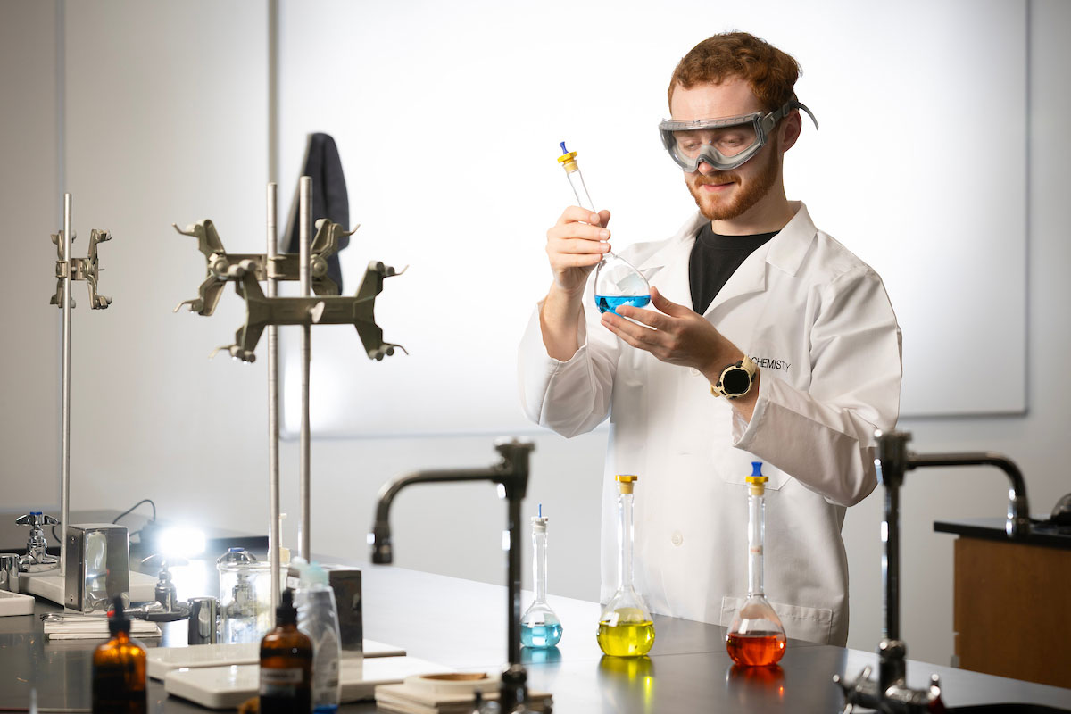 A man stands in a lab setting and holds a glass beaker with a blue substance 