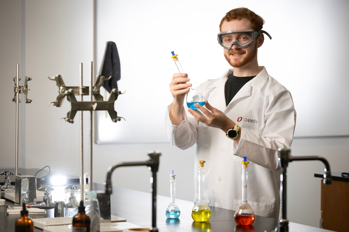 A man stands in a lab setting and holds a glass beaker with a green substance 