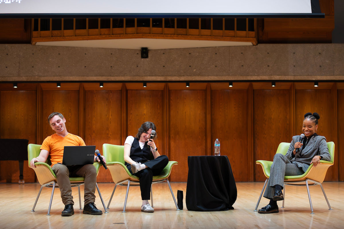Three people sit in chairs on a stage, holding microphones