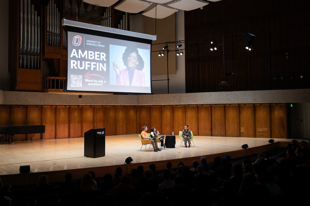 Three people sit in chairs on a stage in front of a full audience, with a projector screen behind them with a photo of Amber Ruffin on it.