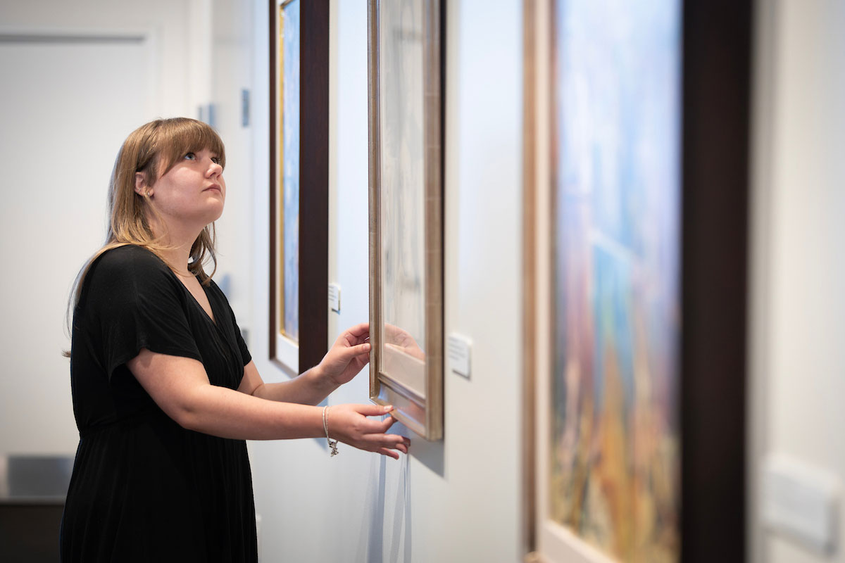 A woman in an art gallery carefully adjusts a painting hanging on a wall.