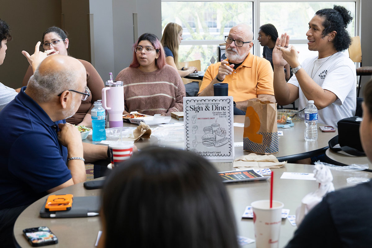 A group of students and faculty sit at table and communicates through sign language.  