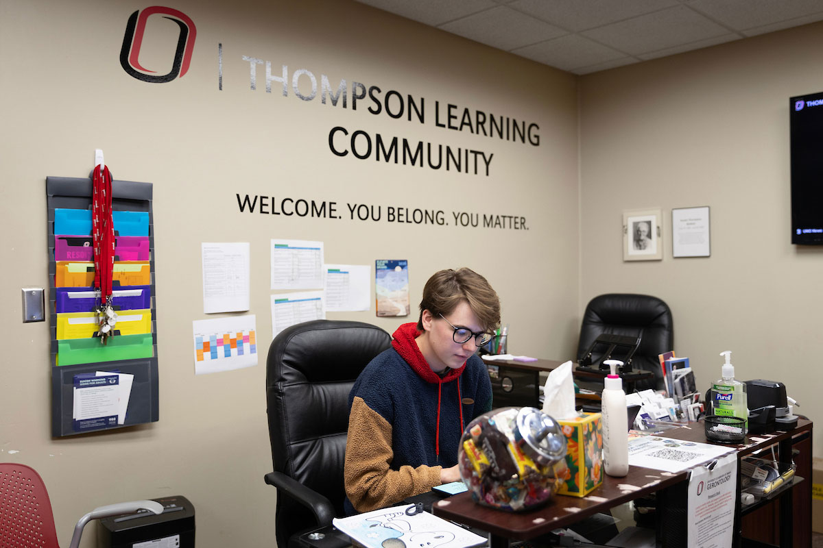 A student sits at a desk working in front of a wall that reads "Thompson Learning Community"