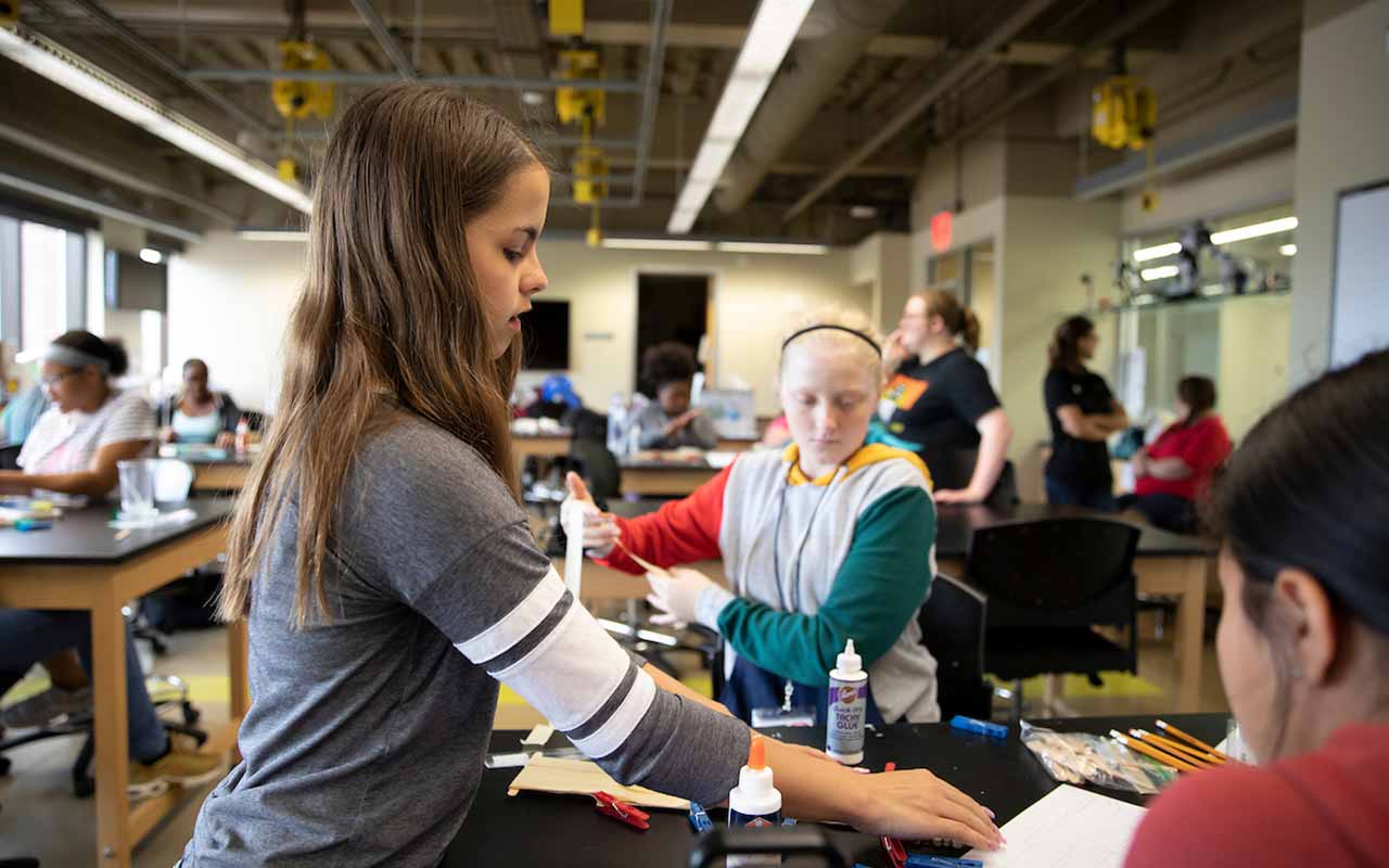 A young female student stands at a desk in a science lab, reaching for materials while speaking to her fellow classmates. 