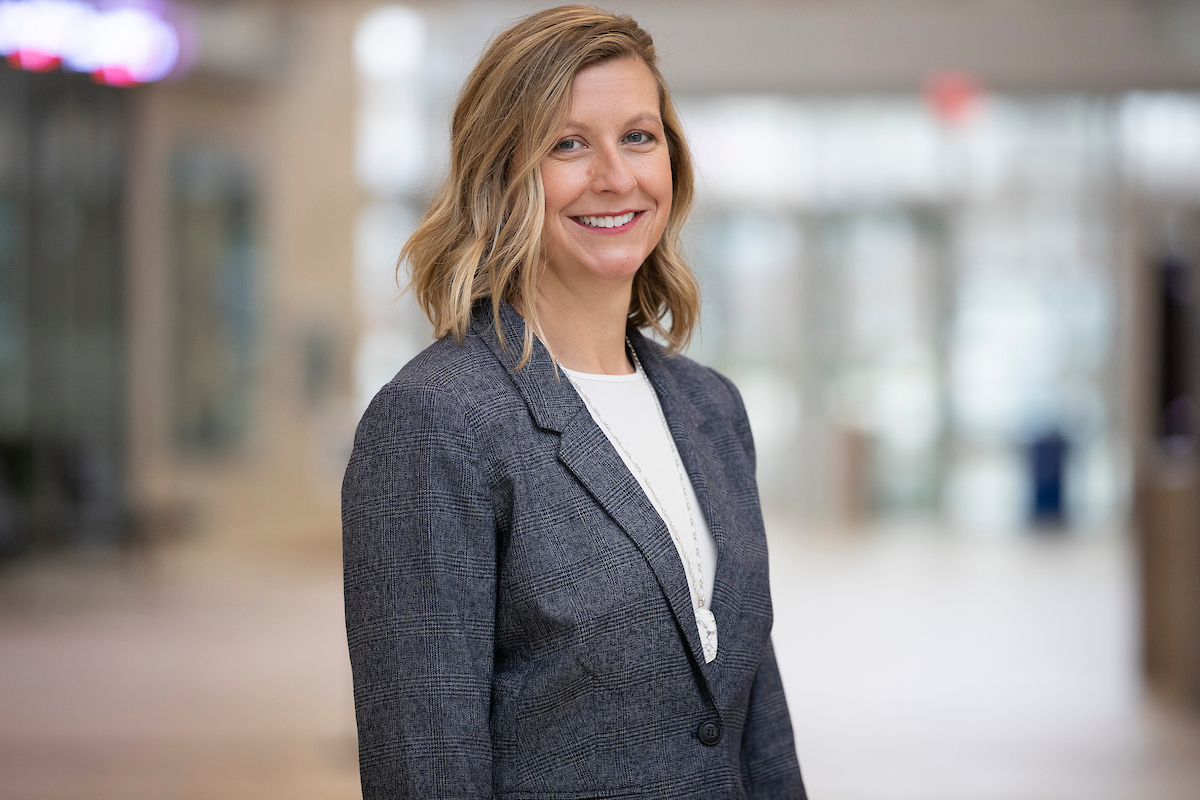 Woman in a gray blazer and white top smiles at the camera in a softly lit indoor hallway setting.