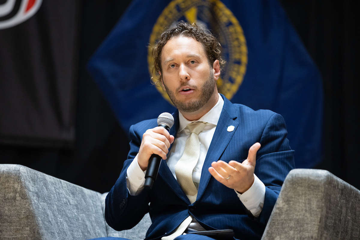 Man in a blue suit and light-colored tie speaks into a microphone while gesturing with his hand, seated on stage