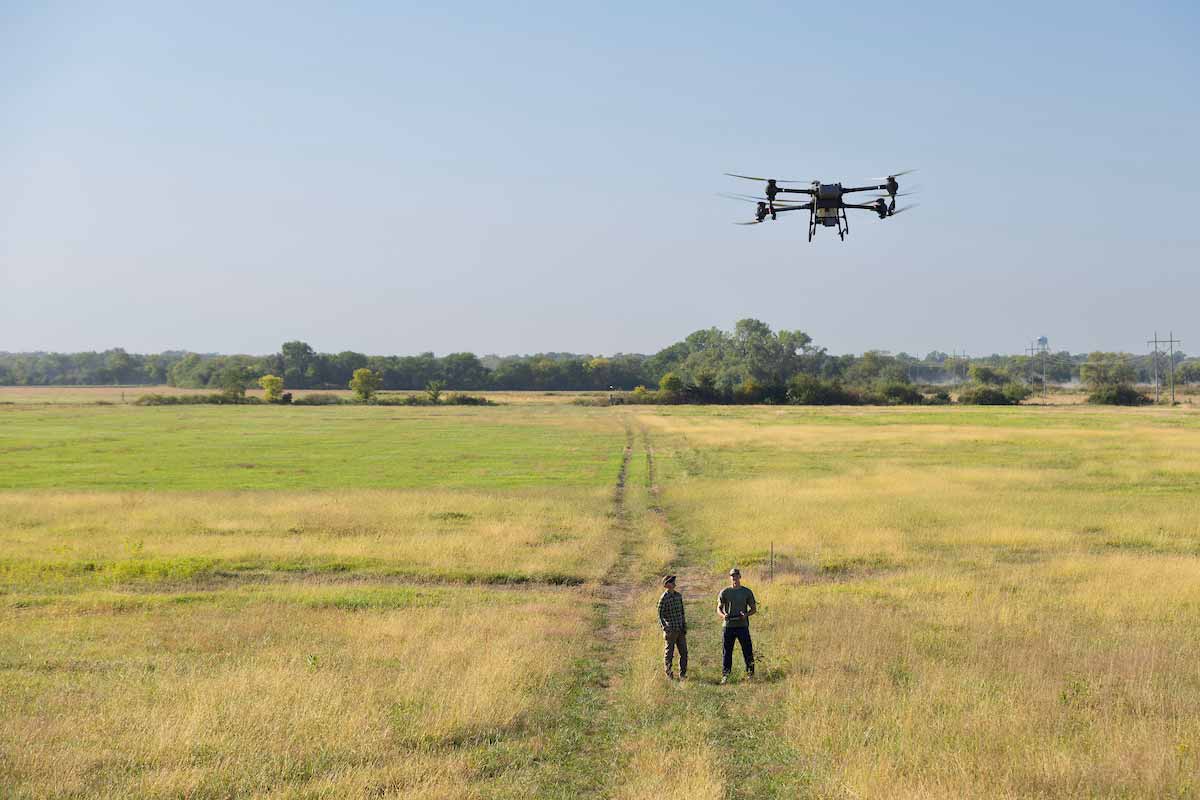 A drone hovers above a wide grassy field while two people stand below on a narrow path, looking up at it under a clear sky.