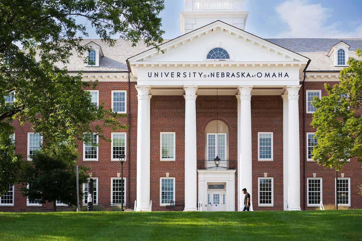 Front view of a red-brick Arts and Sciences Hall at the University of Nebraska at Omaha, featuring tall white columns, a central entrance, and the university’s name displayed above, with green lawn and trees in the foreground and a person walking nearby. 