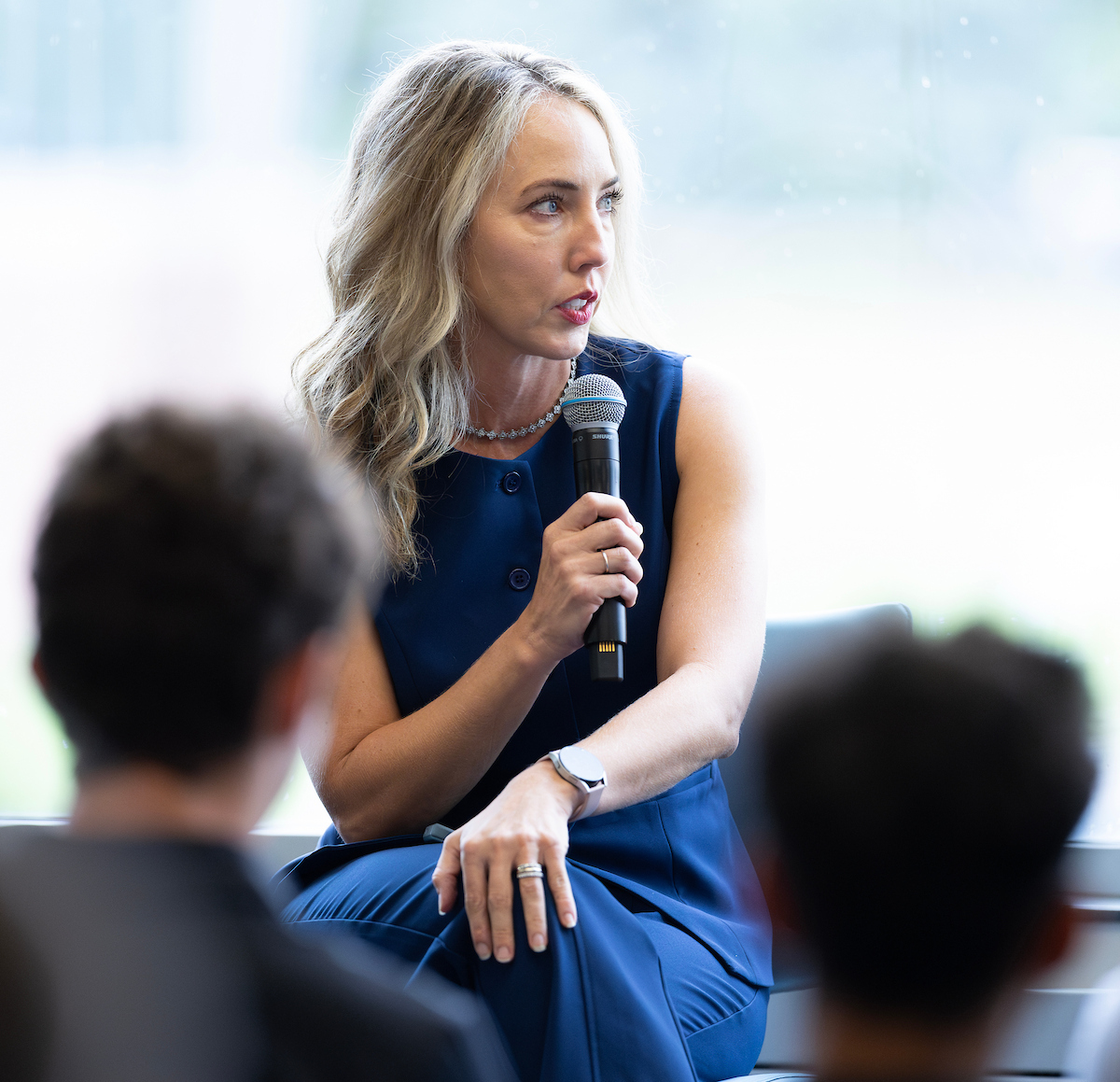 Woman in a blue sleeveless outfit speaks into a handheld microphone while seated, turned slightly to the side, addressing an audience during a panel discussion.