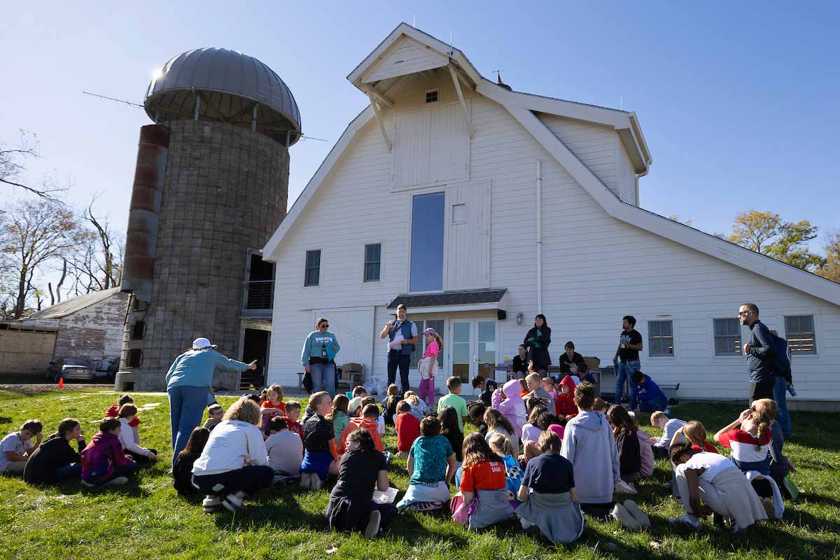 A group of elementary school students sit on the ground with their backs facing the camera, listening to a professor speak with a large white barn and gray silo behind him. 