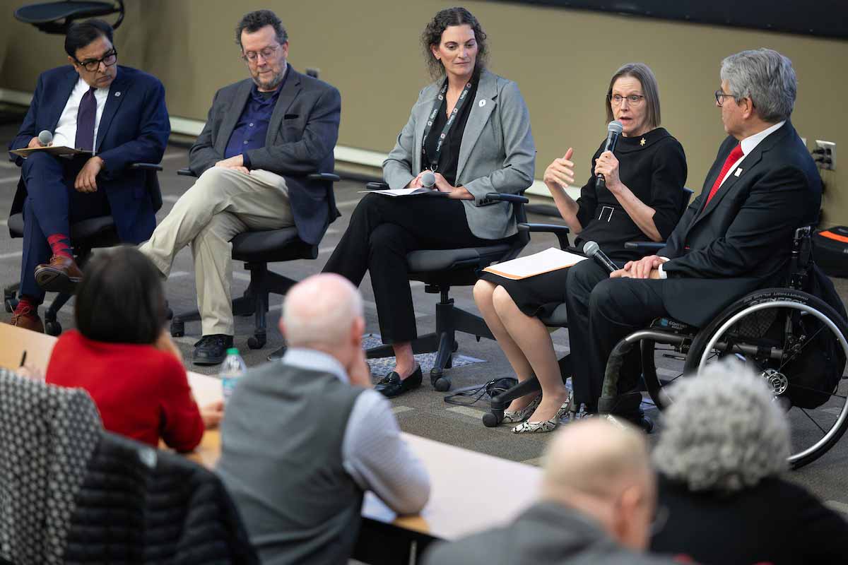 Panel of five speakers seated at the front of a lecture hall, with one woman speaking into a microphone while audience members listen during UNO’s Student Research and Creative Activity Fair. 