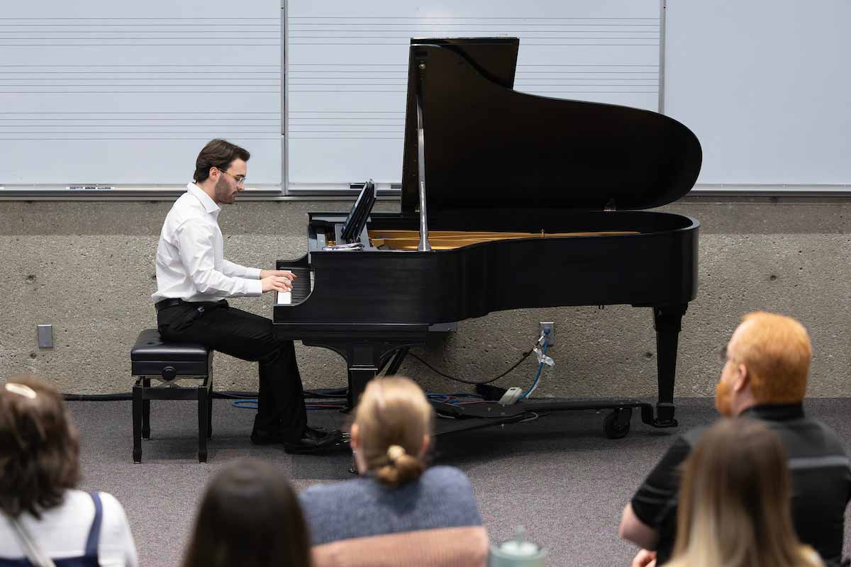 Student in a white shirt plays a grand piano in front of an audience seated in a classroom at UNO’s Student Research and Creative Activity Fair. 