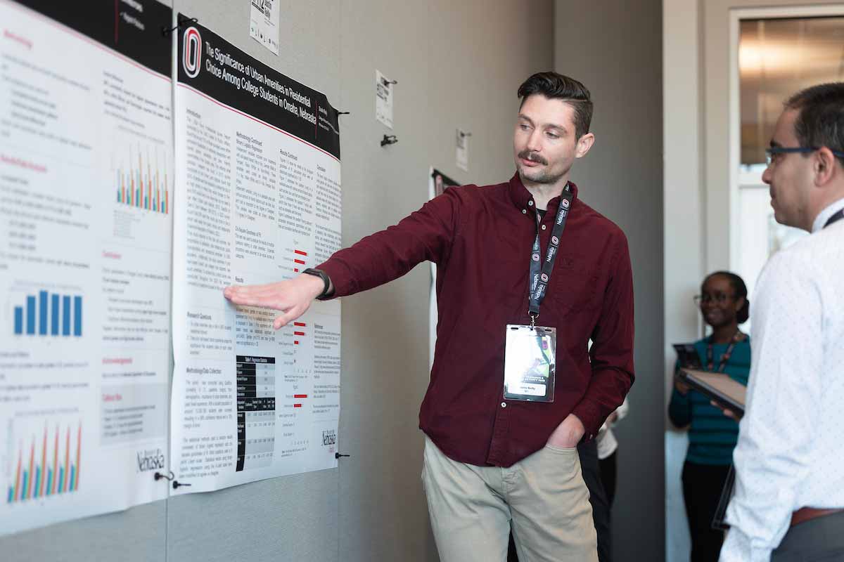 Student presenter gestures toward charts and data on a research poster while explaining findings to an attendee at UNO’s Student Research and Creative Activity Fair. 