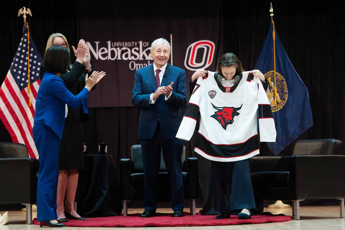 Former U.S. Director of National Intelligence Avril Haines holds up a University of Nebraska at Omaha Mavericks hockey jersey on stage as Chuck Hagel and others applaud during the Hagel Forum in Global Leadership. 