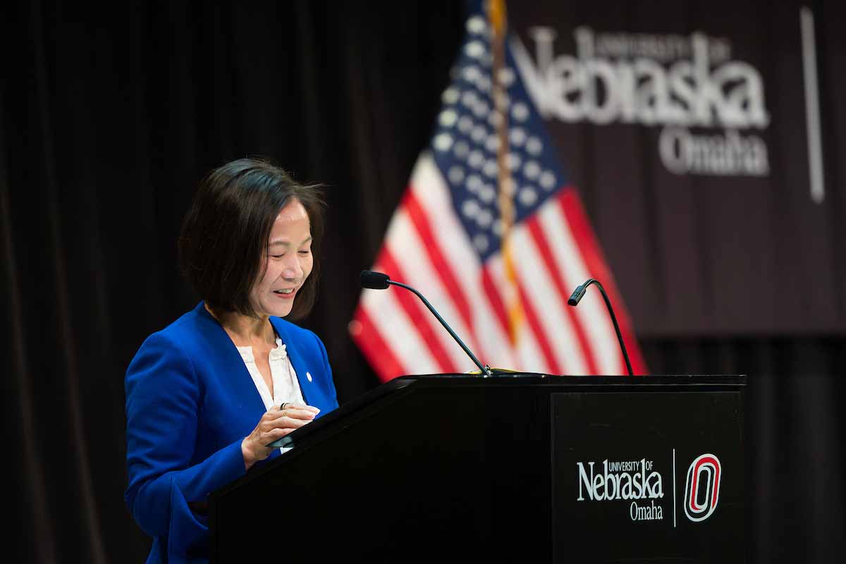 Chancellor Joanne Li speaks at a podium on stage at the University of Nebraska at Omaha during the Hagel Forum in Global Leadership, with an American flag in the background. 