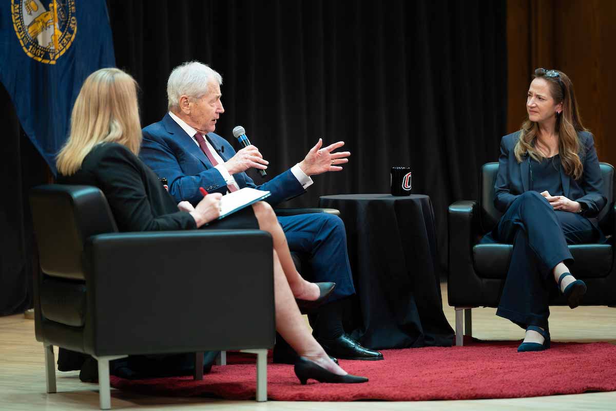 Chuck Hagel speaks into a handheld microphone while seated on stage with Avril Haines and a moderator during the Hagel Forum in Global Leadership at the University of Nebraska at Omaha. 