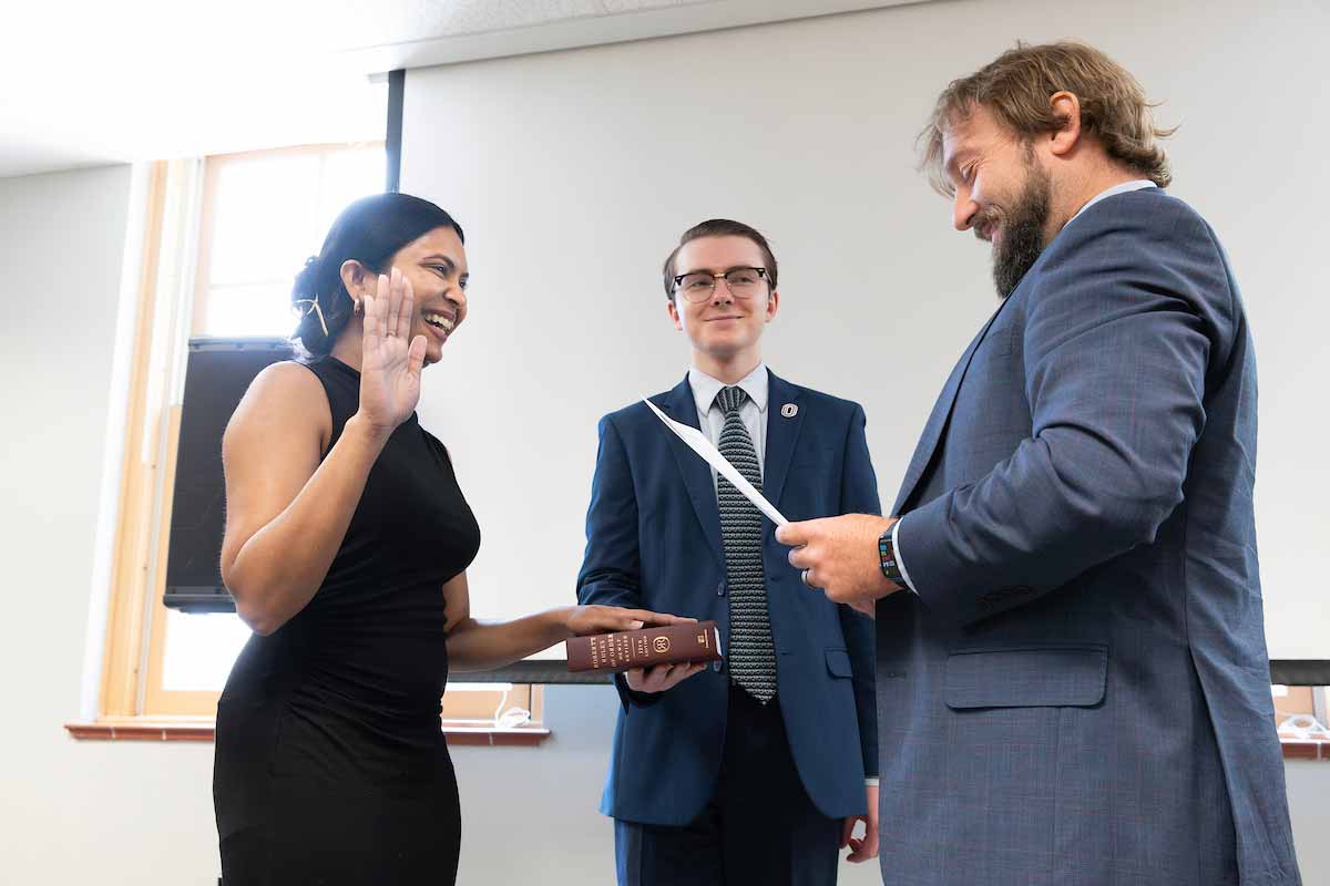 A woman places her left hand on a Robert’s Rules of Order book held by a man while raising her right hand to take an oath, while another man across from her holds a sheet of paper to read her oath.  