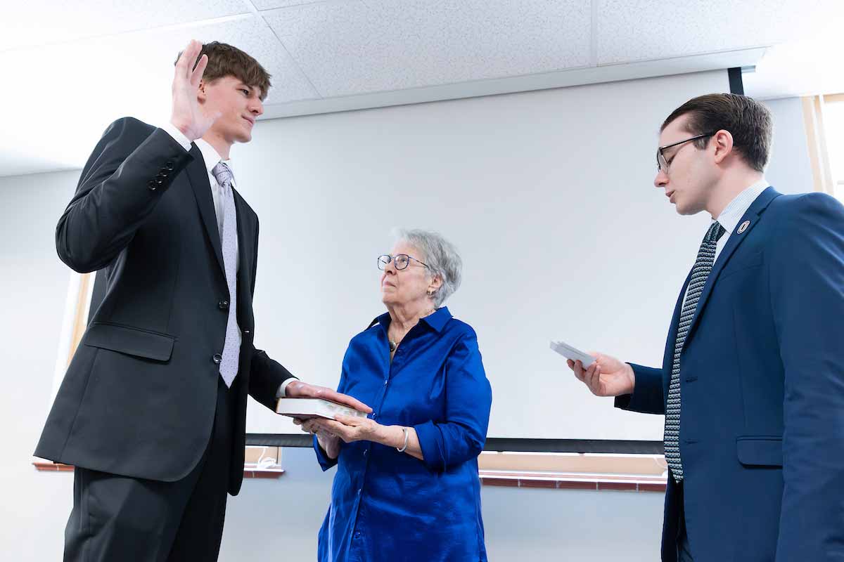 A man places his right hand on a Robert’s Rules of Order book held by a woman while raising his right hand to take an oath, while another man standing across from him holds a piece of paper to read his oath. 
