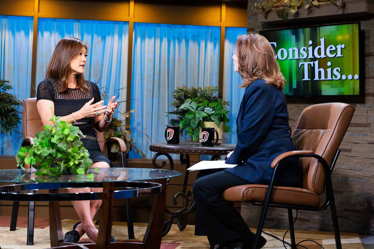 ABC News’ Juju Chang sits in a chair and faces UNO TV’s Cathy Wyatt on a TV set, in front of a screen that reads ‘Consider This,’ 