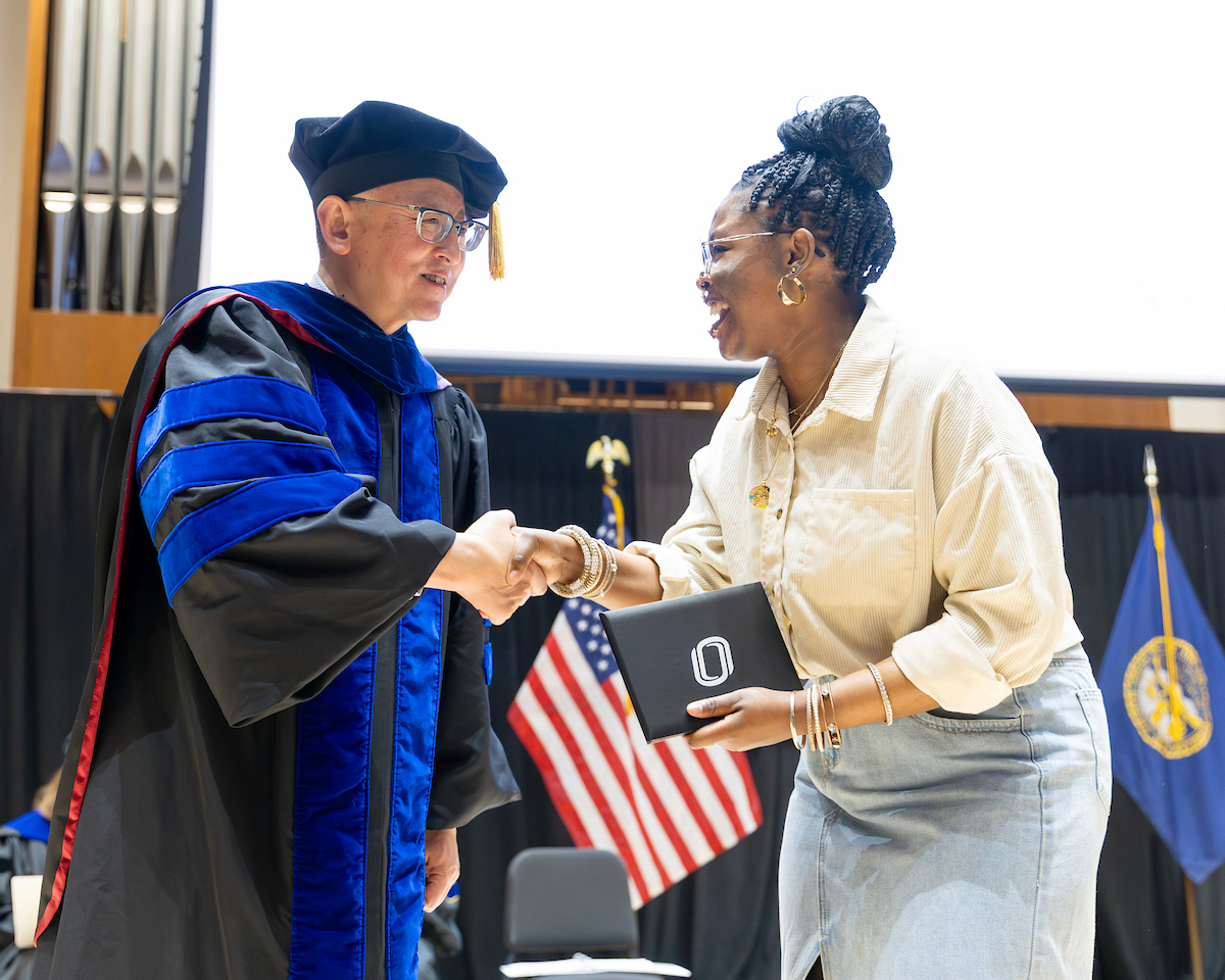 A university official in academic regalia shakes hands with a smiling student holding a UNO-branded award folder on stage during a formal honors ceremony, with U.S. and Nebraska flags in the background. 