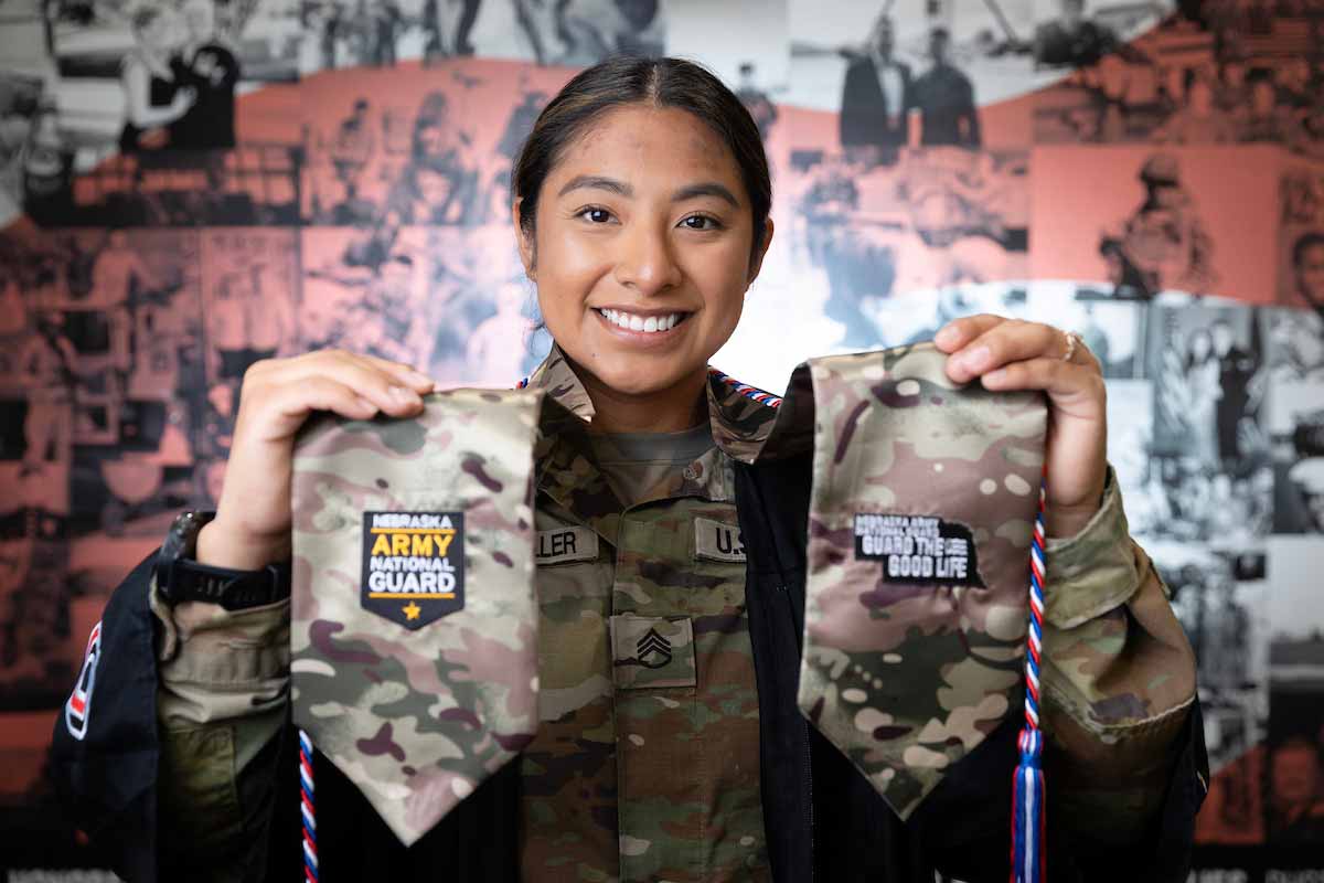 UNO student Blanca Brownmiller smiles as she poses in her graduation gown, holding up her Army National Guard graduation stole in front of a backdrop of military photos. 
