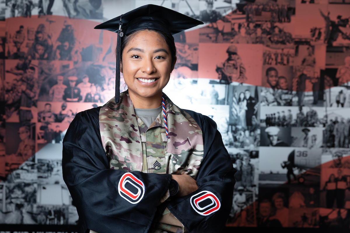 UNO student Blanca Brownmiller smiles as she poses in her graduation gown, holding up her Army National Guard graduation stole in front of a backdrop of military photos. 