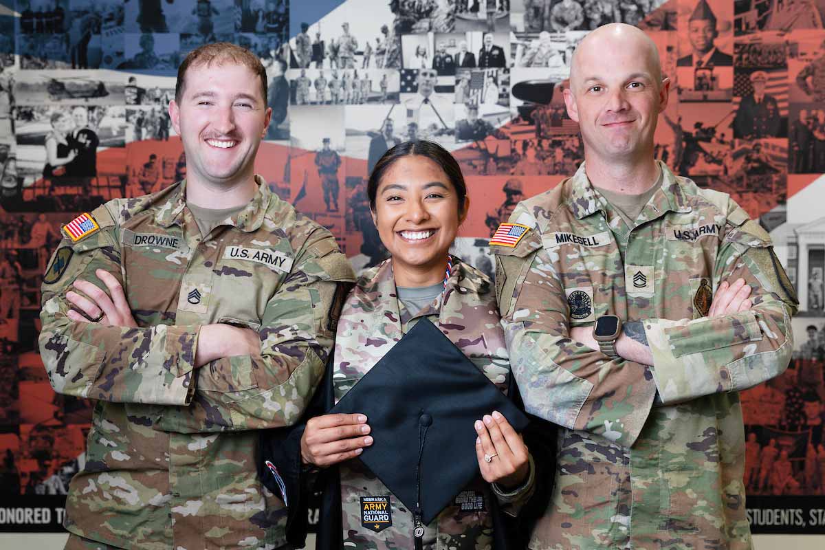 UNO student Blanca Brownmiller smiles as she poses in her graduation gown worn over her Nebraska Army National Guard uniform, while holding her graduation cap. She stands between two fellow soldiers who are smiling with their arms crossed, wearing their Nebraska Army National Guard uniforms in front of a backdrop of military photos.