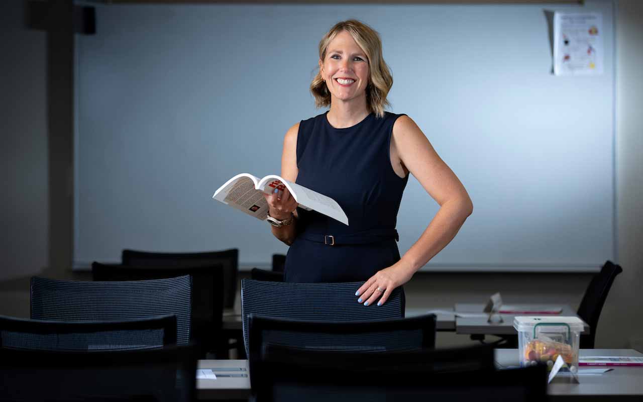 UNO's Jennifer Lemke stands in a classroom holding an open book and smiling toward the camera.