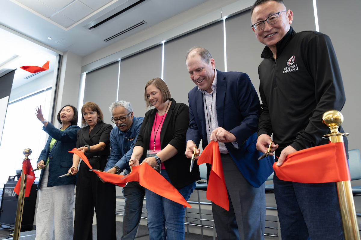 Six people stand in a row at a ribbon-cutting ceremony, smiling as they use scissors to cut a red ribbon stretched between gold stanchions. 