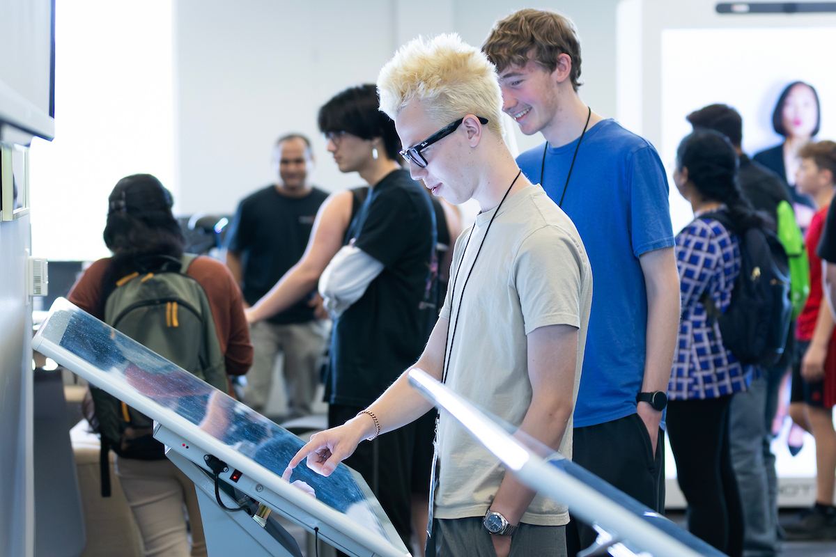 Students interact with a large touchscreen display. In the foreground, a student with light blond hair and glasses uses the screen while another student stands nearby watching.