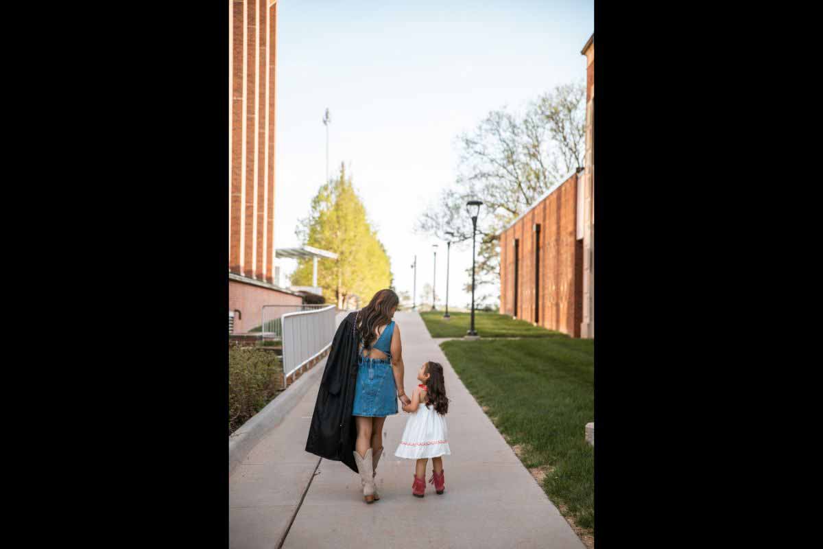 UNO graduate Blacna Brownmiller walks on campus with her back to the camera, holding her graduation regalia over her shoulder while holding hands and looking down at her daughter. 