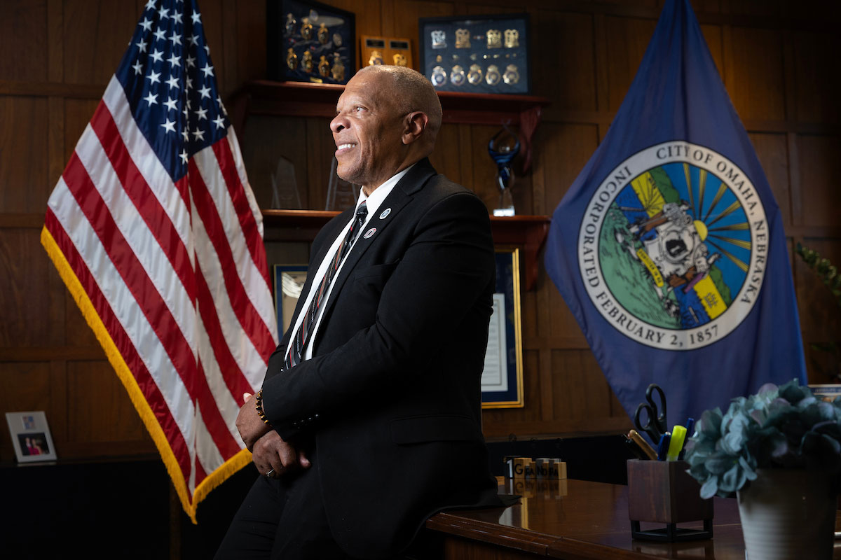 A man sits on the edge of his desk, looking away from the camera.