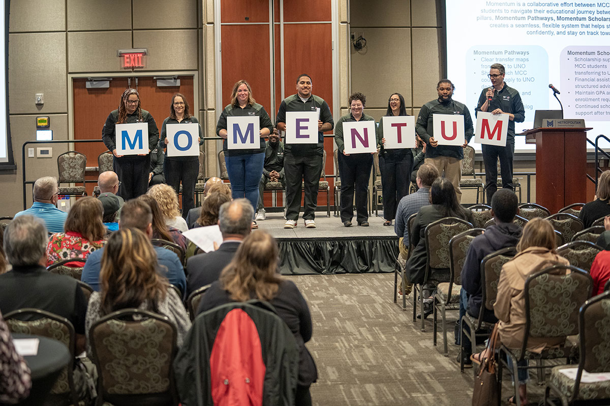 A group of 8 MCC and UNO employees stand on a stage and hold signs that spell 'Momentum'