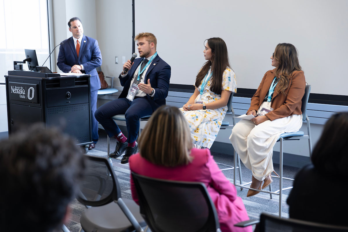 A panel discussion takes place at OMAXAI where three speakers sit on stools while one panelist speaks into a microphone. A moderator stands at a podium, and audience members are seated in the foreground listening to the discussion.
