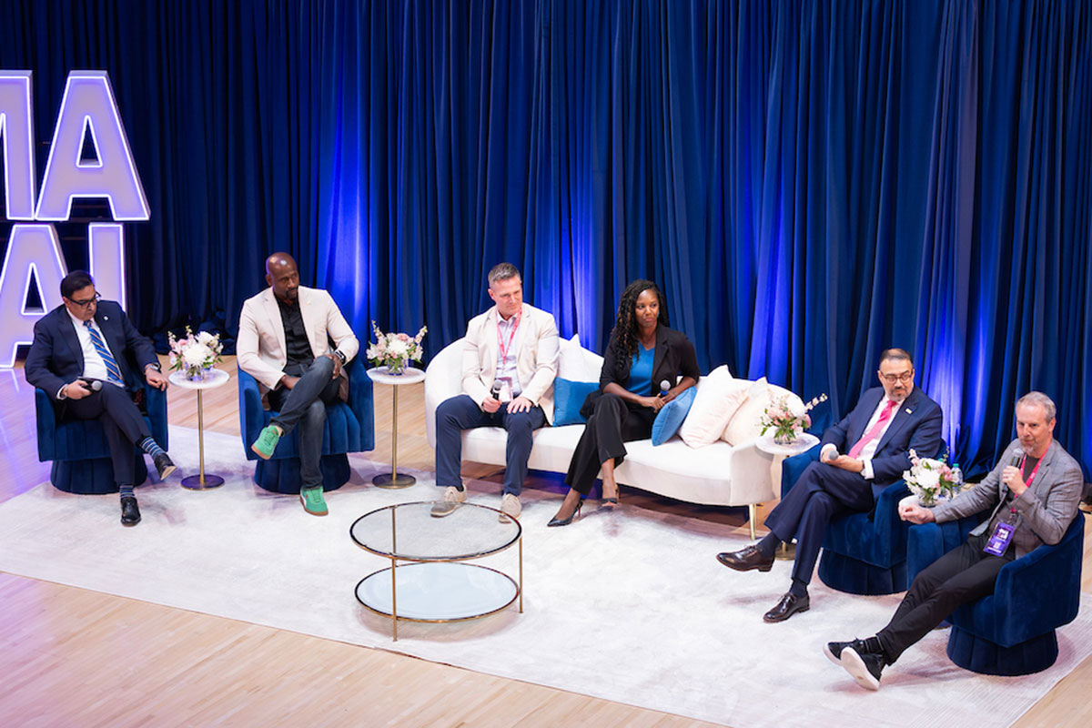 Five panelists and a moderator sit on stage in blue chairs and a white sofa during the OMAXAI conference discussion, with one speaker holding a microphone. Blue drapes, floral arrangements, and large illuminated "OMAHAXAI" letters create a professional event backdrop.