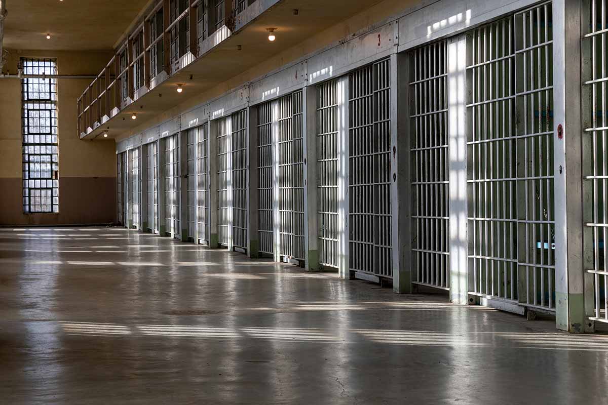 A two-story cell block inside a prison, with rows of barred cells illuminated by sunlight streaming through large windows. 