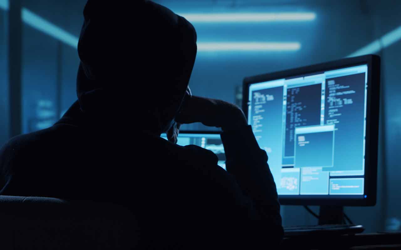 A silhouetted person wearing a hooded sweatshirt sits at a computer workstation in a dimly lit room, facing multiple monitors displaying lines of code and software windows, creating a cybersecurity or hacking-themed scene. 
