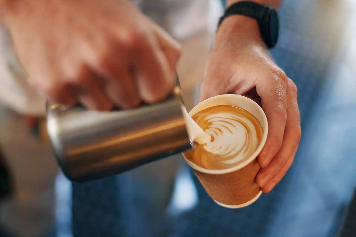Latte art, coffee shop and hands pouring cup for take away with barista, small business and hospitality. Cappuccino, milk foam and service person in cafe, restaurant or bistro with hot drink to go. 