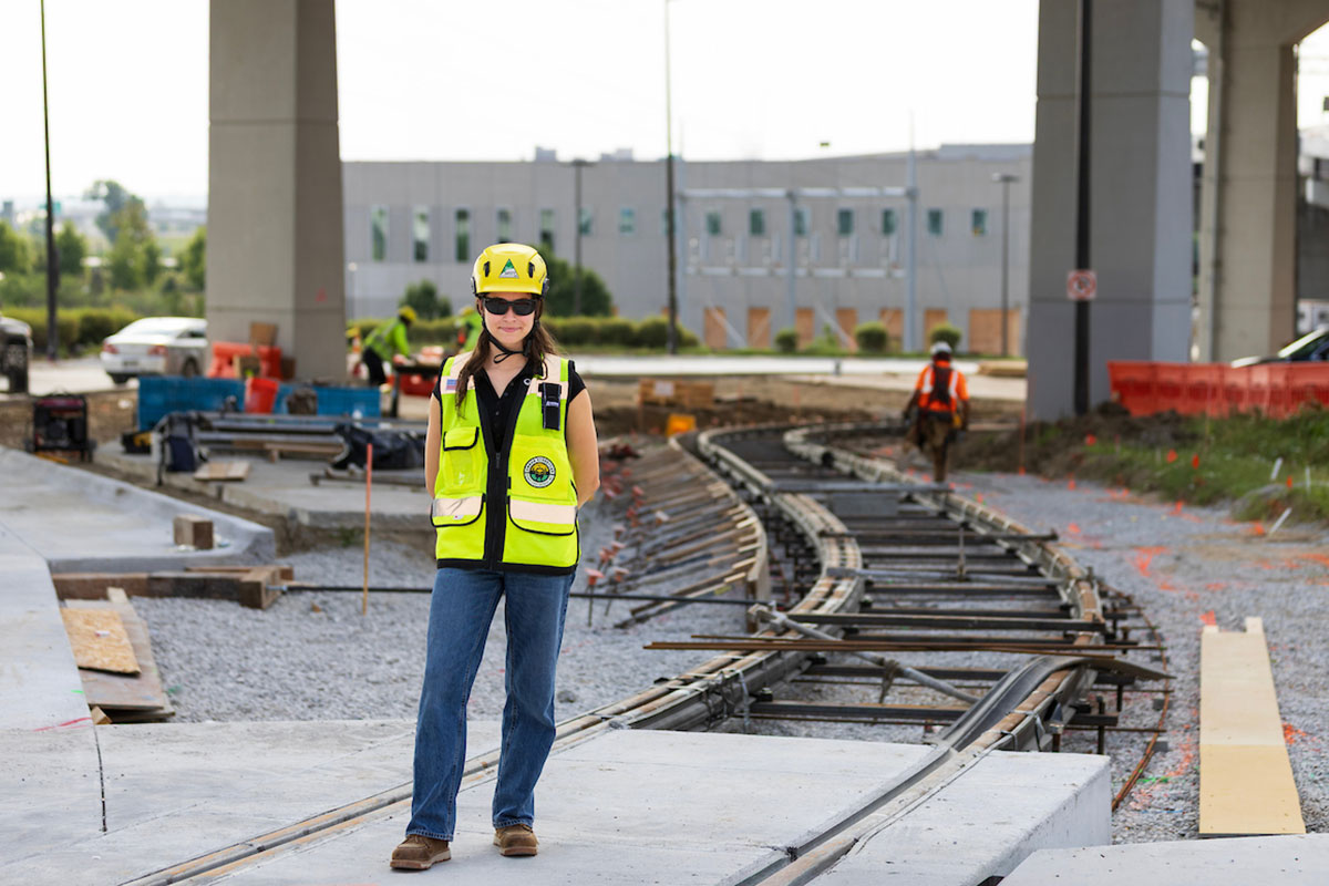 A woman in a refelctive vest and a hard hat stands with her arms crossed on the downtown Omaha streetcar tracks in front of the Mutual of Omaha building.