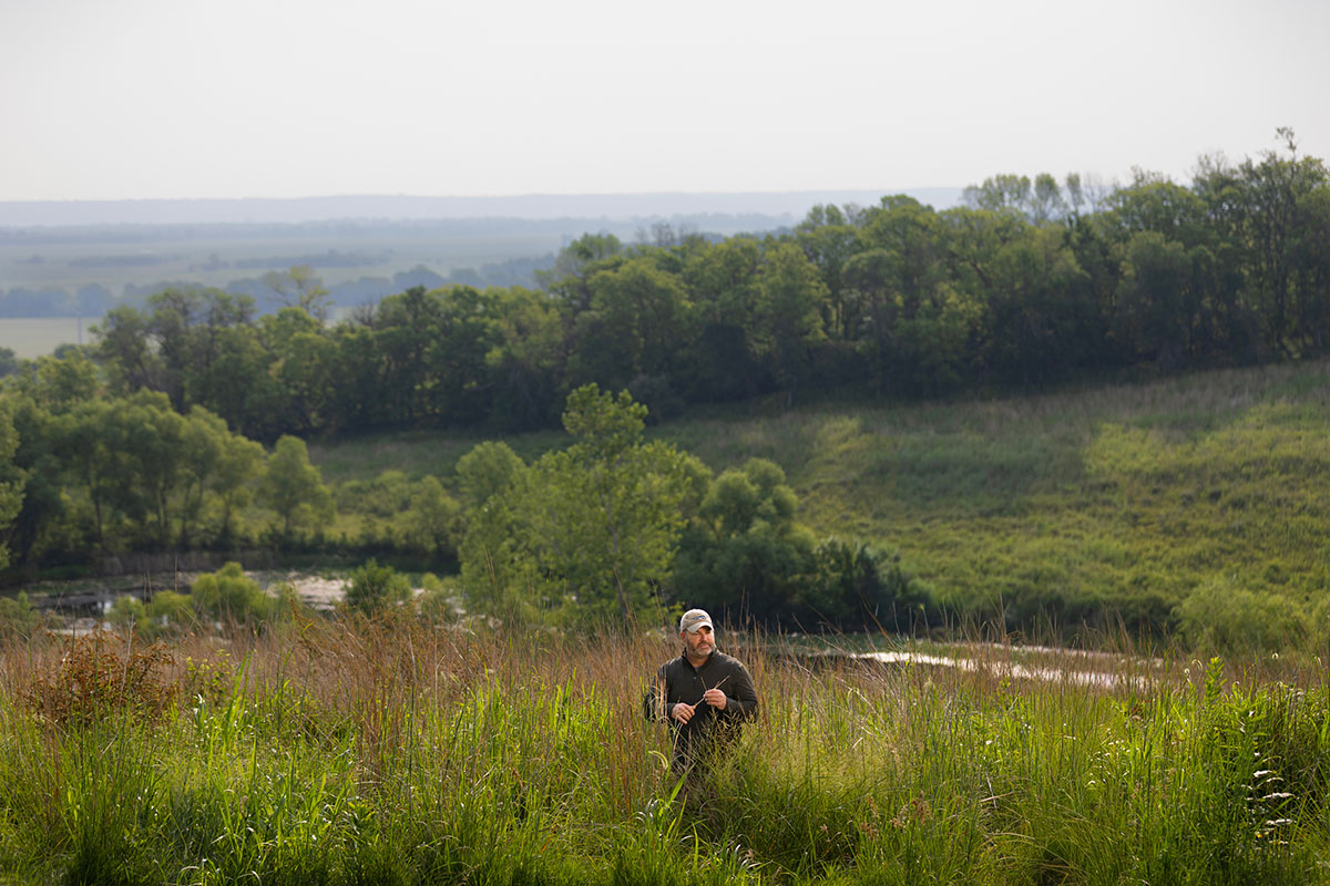A man in a black long sleeve shirt and tan baseball cap stands in the middle of a prairie with a small lake and tree-covered hill behind him