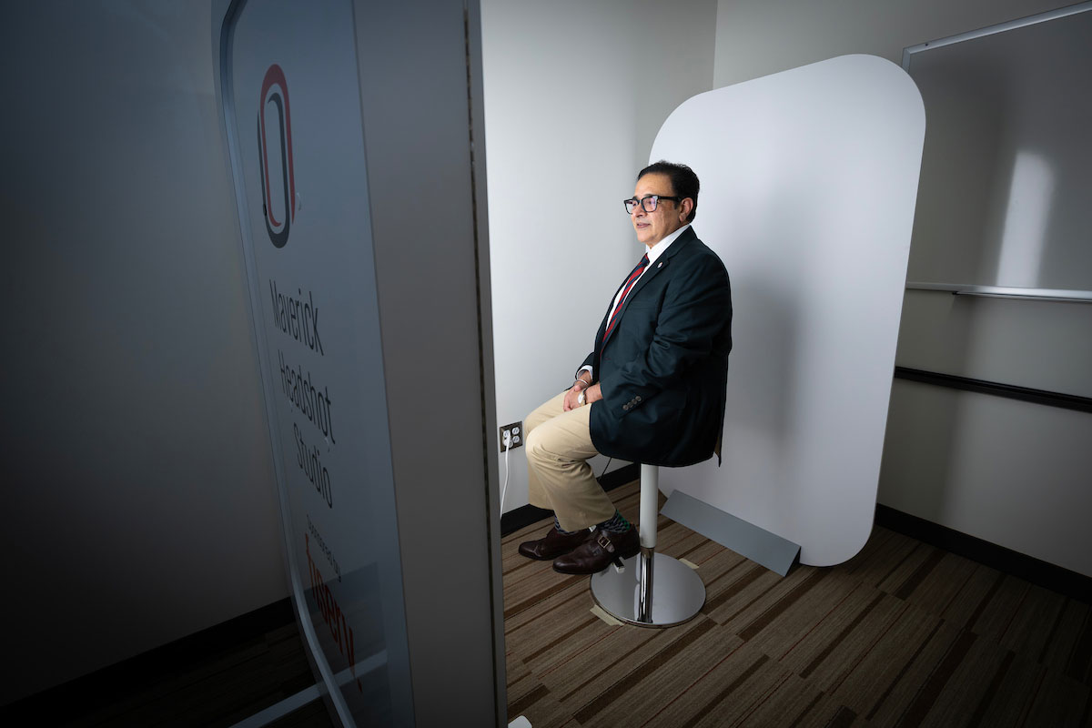 A man sits in a chair to have his headshot taken by an automated machine, the Maverick Headshot Studio