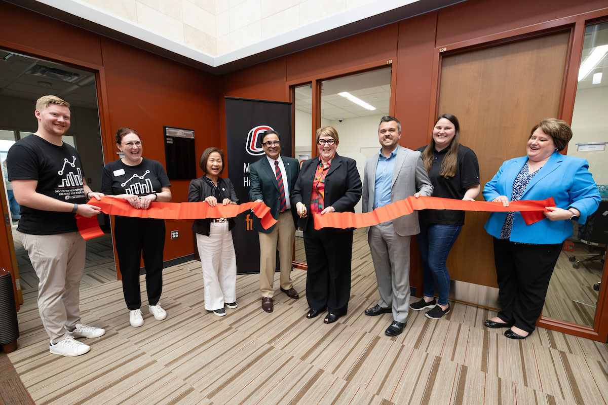 A group of 8 people hold a large red ribbon that has been cut to celebrate the opening of the Maverick Headshot Studio
