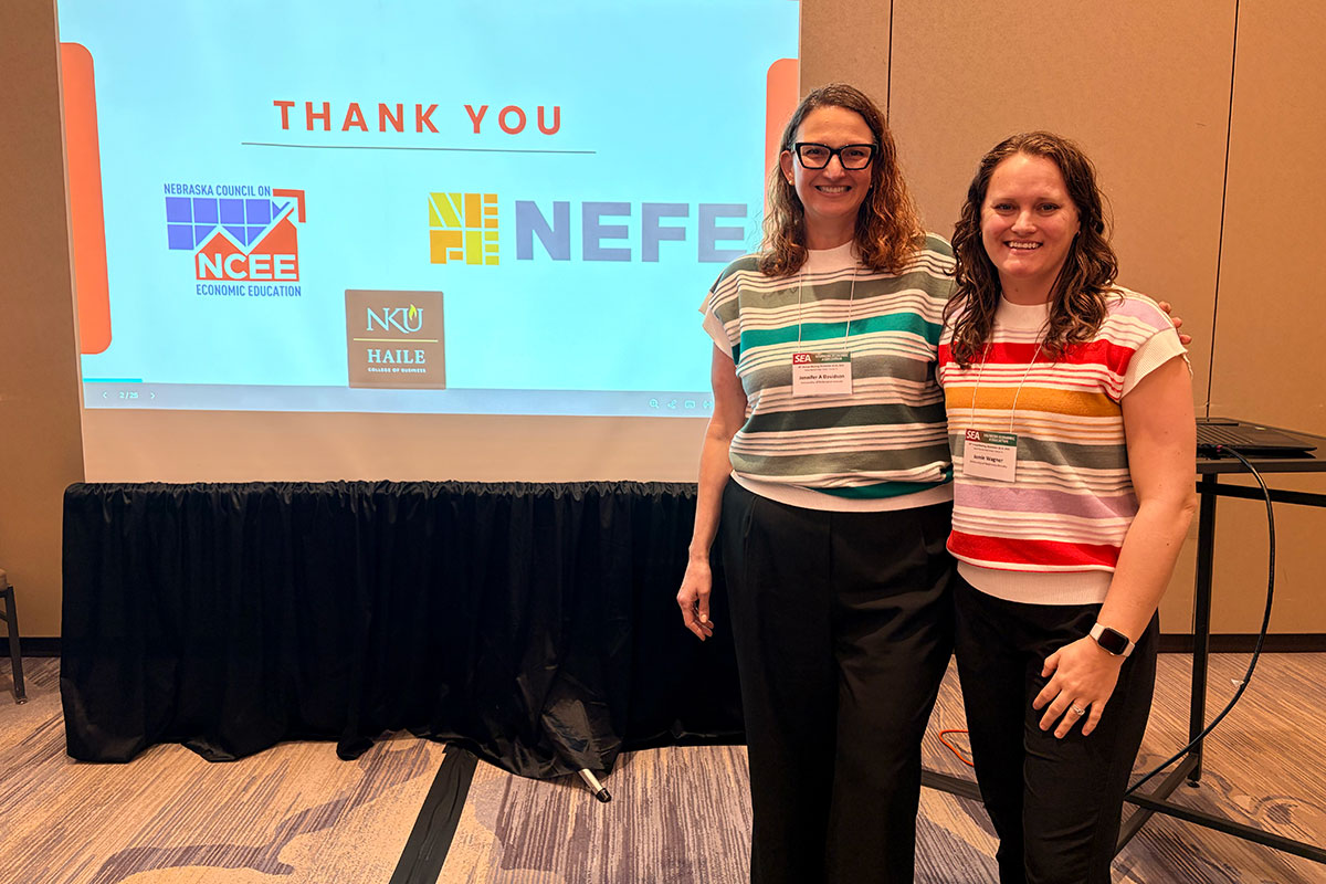 Two women wearing identification lanyars stand side by side in front of a projector that reads "Thank you, Nebraska Council on Economic Education, NEFE"