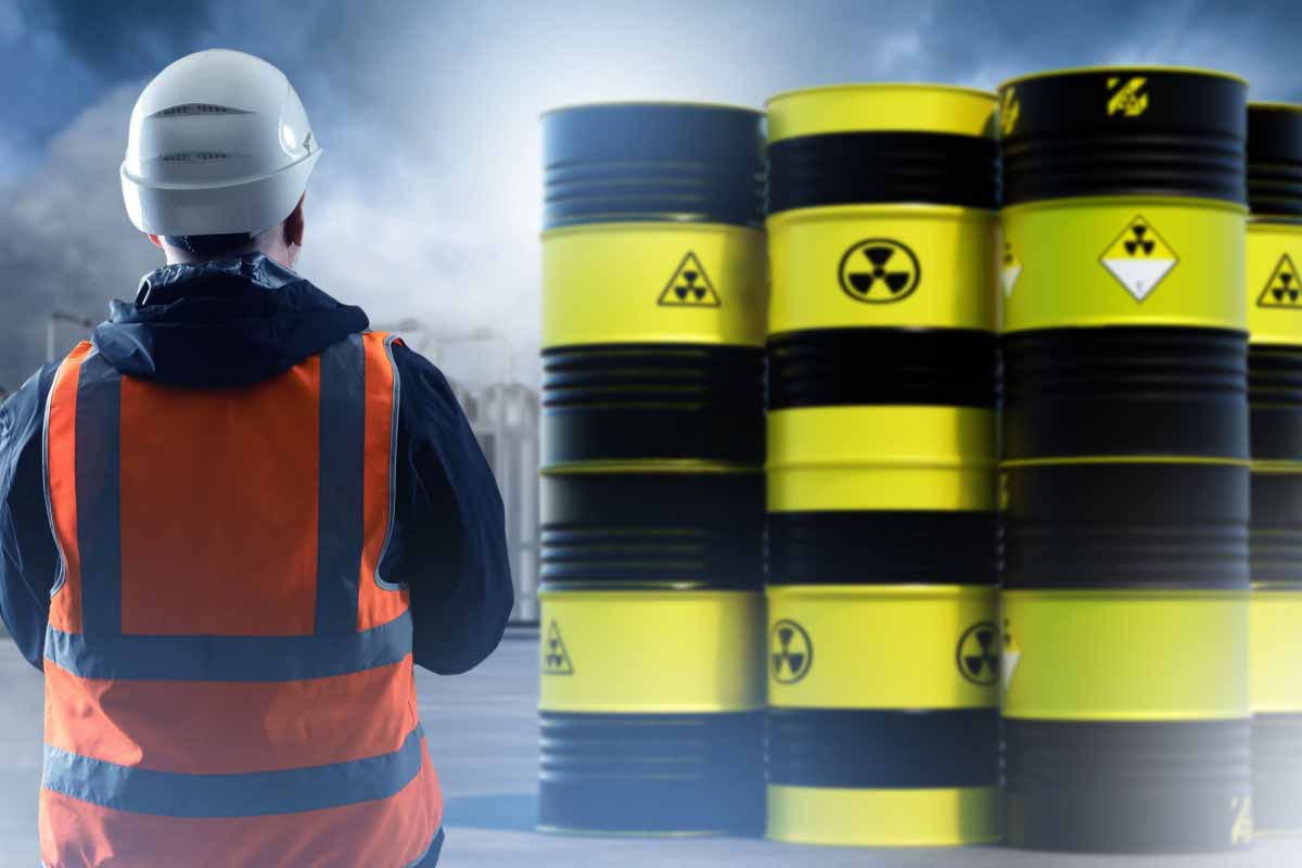 A person in safety gear facing stacked yellow and black nuclear waste barrels at an industrial site under a cloudy sky. 