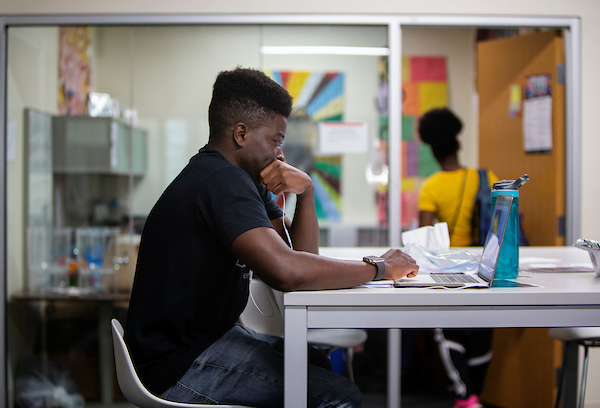 a young black man sits at a desk looking at a laptop.