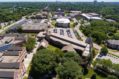 Aerial view of a large college campus with multiple academic buildings, parking lots, and tree-filled green spaces stretching into the distance.