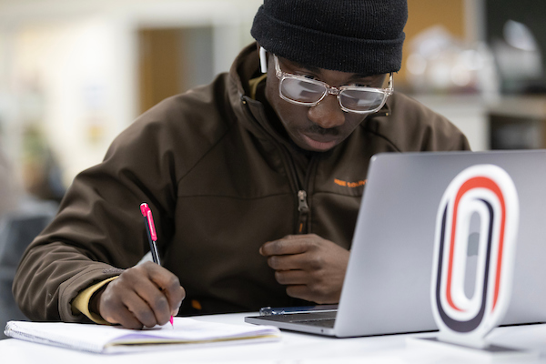 a black man wearing glasses and wearing a green hoodie writes in a notebook as he works on his laptop.